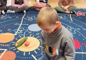 Dzieci siedzą na dywanie podczas zajęć. Jeden z chłopców ogląda znajdujące się w misce warzywa.