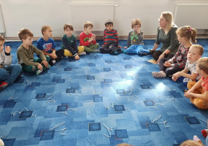 Dzieci wraz z Panią bibliotekarką siedzą w kręgu na dywanie.