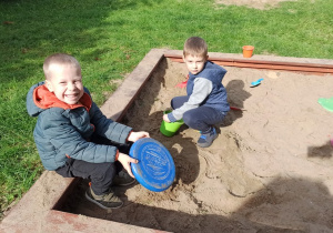 Dwóch chłopców bawi się w piaskownicy. Do zabawy wykorzystują wiaderko i łopatki.
