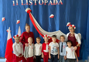 grupa dzieci ze swoimi paniami pozują do zdjęcia na tle patriotycznej dekoracji
