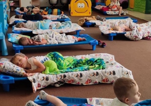 Dzieci śpią na leżakach, ustawionych na dywanie w sali przedszkolnej.