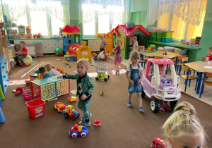 Dzieci bawią się w sali przedszkolnej. Na dywanie znajdują się zabawki: samochody, klocki, wózki, piłki. Przedszkolaki wykorzystują je do zabawy.