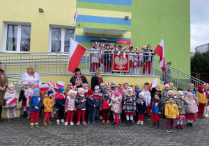 Grupa dzieci i nauczycielki stoją przed budynkiem przedszkola. Przygotowują się do uroczystego zaśpiewania Mazurka Dąbrowskiego, w tle flagi i godło.