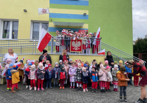 Grupa dzieci i nauczycielki stoją przed budynkiem przedszkola. Przygotowują się do uroczystego zaśpiewania Mazurka Dąbrowskiego, w tle flagi i godło.