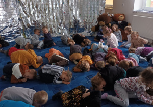 grupa dzieci uczestniczy w zajęciach, leżą na dywanie, na pluszowych misiach