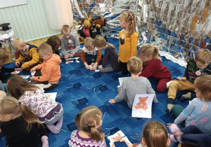 grupa dzieci siedzi na dywanie w bibliotece , w małych podgrupach wykonują rózne zadania