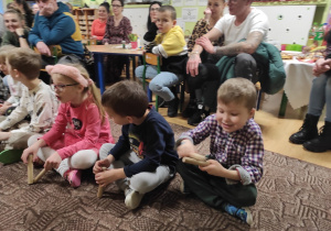 dzieci siedzą na dywanie, przed nimi leżą instrumenty, obok siedzą rodzice