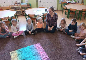 grupa dzieci i nauczycielka siedzą na dywanie, biorą udział w zabawie edukacyjnej
