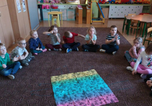 grupa dzieci siedzi na dywanie, biorą udział w zabawie edukacyjnej, na środku leży kolorowa chustka, pod którą ukryte są pomoce dydaktyczne