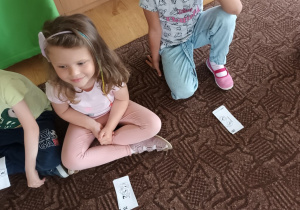Dzieci siedzą na dywanie. Każdy z przedszkolaków ma przydzielony zestaw karteczek, imitujących pieniądze.