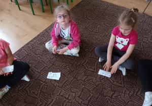 Dzieci siedzą na dywanie. Każdy z przedszkolaków ma przydzielony zestaw karteczek, imitujących pieniądze.