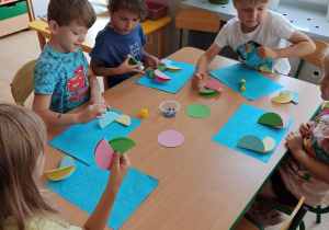 grupa dzieci przy stoliku wykonuje pracę plastyczną na niebieskich kartkach