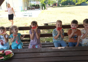 grupa dzieci siedzi w altanie w przedszkolnym ogrodzie. Wszyscy jedzą arbuzy