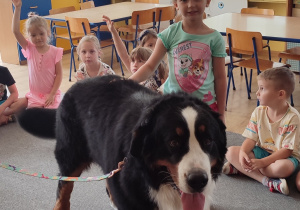 na środku klasy na dywanie stoi czarny pies, obok niego dziewczynka, w tle dzieci