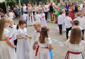 grupa dzieci tańczy walca , dziewczynki w białych sukienkach, chłopcy ubrani na galowo, w tle rodzice