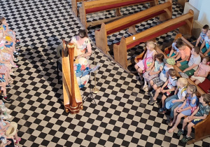 wnętrze kościoła, artystka gra na harfie, wokół niej zgromadzone dzieci