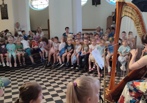 wnętrze kościoła, artystka gra na harfie, wokół niej zgromadzone dzieci