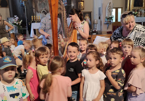 wnętrze kościoła, artystka prezentuje harfę, wokół niej zgromadzone dzieci i nauczyciele, pozują do zdjęcia
