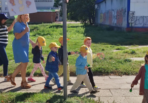 nauczycielki i dzieci maszerują ulicami miasta , dzieci w czapeczkach z logo przedszkola, z emblematami grup, z instrumentami w rękach. Niektóre z nich przebrane w stroje grupowe