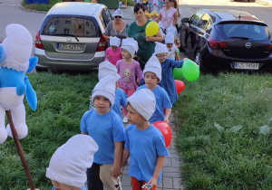 nauczycielki i dzieci maszerują ulicami miasta , dzieci w czapeczkach z logo przedszkola, z emblematami grup, z instrumentami w rękach. Niektóre z nich przebrane w stroje grupowe