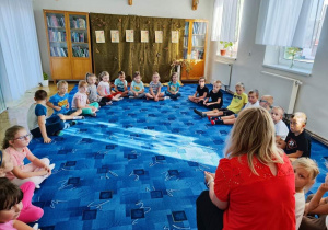 Dzieci z grupy "Biedronki" siedzą w kole na dywanie. Słuchają tego, co mówi do nich Pani bibliotekarka.