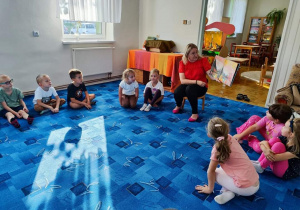 Dzieci z grupy "Biedronki" siedzą w kole na dywanie. Słuchają opowiadania, które czyta Pani bibliotekarka.