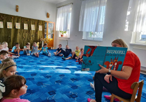 Dzieci z grupy "Biedronki" siedzą w kole na dywanie. Słuchają opowiadania, które czyta Pani bibliotekarka.