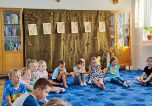 Dzieci z grupy "Biedronki" siedzą w kole na dywanie. Słuchają opowiadania, które czyta Pani bibliotekarka. W tle znajduje się jesienna dekoracja.