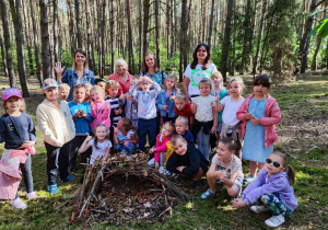 Dzieci z grupy Biedronki wraz z nauczycielkami i Panią botanik pozują do zdjęcia. Przed nimi znajduje się domek dla jeży.
