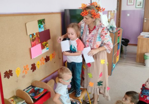 nauczycielka przebrana w jesienny stój prowadzi zajęcie, wokół niej dzieci, jedno z nich przypina do tablicy obrazki