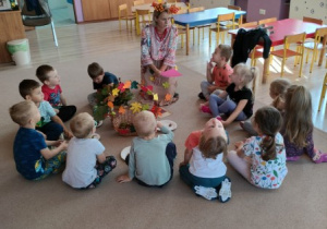 nauczycielka przebrana w jesienny stój prowadzi zajęcie, wokół niej na dywanie siedzą dzieci