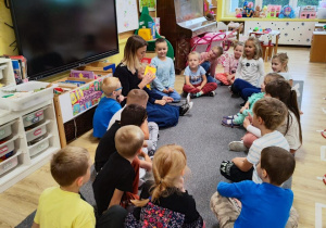 Przedszkolaki wraz z nauczycielką siedzą na dywanie. Nauczycielka wskazuje kolejne strony książki. Wszyscy prowadzą rozmowę.