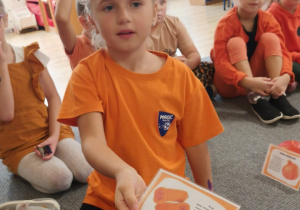 Dzieci siedzą w kole na dywanie, ubrane są w pomarańczowe stroje. Jedna z dziewczynek trzyma obrazek, przekazany jej przez nauczycielkę.