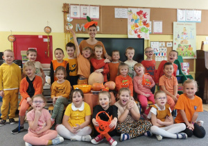 Dzieci wraz z nauczycielką pozują do zdjęcia. Wszyscy ubrani są w pomarańczowe stroje.
