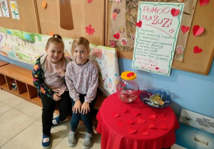 dwie dziewczynki siedzą na ławce w szatni, obok nich na stoliku jest skarbonka i plakat z informacją o akcji charytatywnej