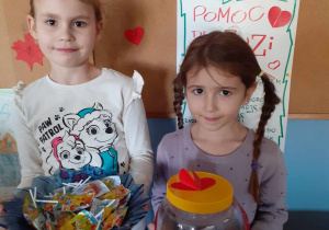 dwie dziewczynki stoją obok plakatu z informacją o akcji dla Zuzi, jedna trzyma miskę z lizakami, drga skarbonkę z pieniędzmi