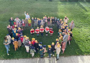 społeczność przedszkolna na podwórku , osoby ustawione w kształt serca. w jego środku stoi 5 dzieci , które trzymają serca z literkami układającymi się w imię ZUZIA