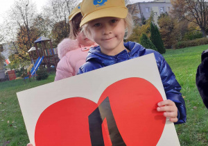 dziewczynka na podwórku przedszkolnym, w dłoniach trzyma serce z literką A