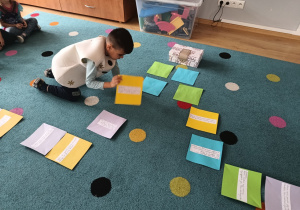 chłopiec kuca na dywanie, wokół niego rozłożone kolorowe kartki z zadaniami