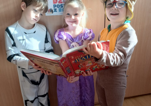 troje dzieci w przebraniach pozuje do zdjęcia z książką w dłoni