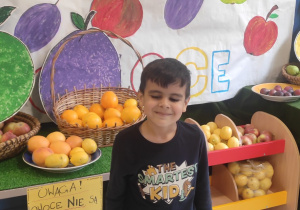 chłopiec pozuje do zdjęcia na tle owocowego stoiska przygotowanego w przedszkolnej szatni , za nim napis RECYKLING DAJE OWOCE