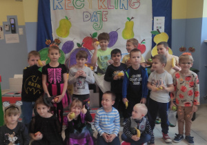 grupa dzieci pozuje do zdjęcia na tle owocowego stoiska przygotowanego w przedszkolnej szatni , za nim napis RECYKLING DAJE OWOCE