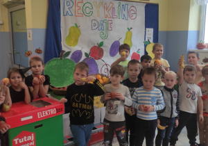 grupa dzieci pozuje do zdjęcia na tle owocowego stoiska przygotowanego w przedszkolnej szatni , za nim napis RECYKLING DAJE OWOCE