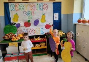 troje dzieci pozuje do zdjęcia na tle napisu Recykling daje owoce. W rączkach trzymają kartonowe duże owoce