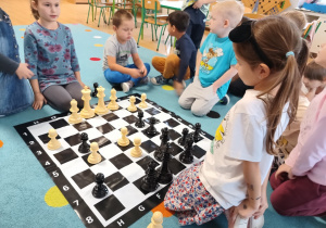 grupa dzieci siedzi na dywanie, na środku leży plansza do gry w szachy i duże pionki
