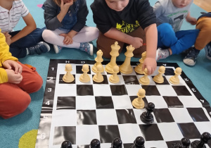 grupa dzieci siedzi na dywanie, na środku leży plansza do gry w szachy i duże pionki, jedno z dzieci wykonuje ruch