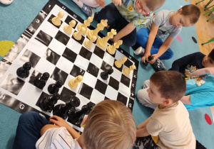 grupa dzieci siedzi na dywanie, na środku leży plansza do gry w szachy i duże pionki
