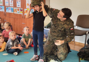 grupa dzieci siedzi na dywanie, umundurowany żołnierz przymierza hełm dziewczynce