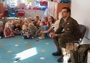 grupa dzieci siedzi na dywanie, umundurowany żołnierz opowiada o swojej profesji