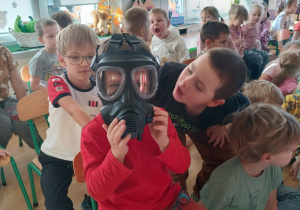 chłopiec przymierza maskę p/gaz, wokół niego zgromadzona duża grupa rówieśników